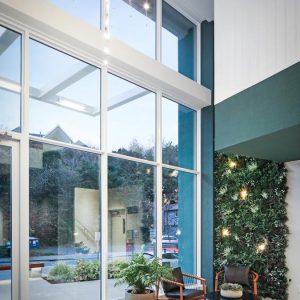 Modern lobby with floor-to-ceiling windows, greenery wall with lights, a potted plant, two chairs, a small table, and a geometric chandelier. Outdoor trees and buildings are visible through the glass.