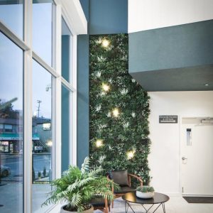 Modern lobby with large windows, a vertical green plant wall with lights, a black chair, a round table, and a potted plant on a terrazzo floor; outside buildings are visible through the glass.