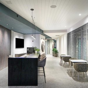 Modern office lounge with a long black counter, bar stools, a TV, potted plants, and small tables with chairs by large windows covered with sheer curtains. Room is well-lit with ceiling lights.