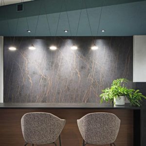 Modern bar area with two upholstered chairs at a wooden counter, a potted green plant on the right side, and three pendant lights illuminating a dark marble-patterned wall in the background.