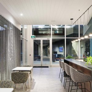 Modern lobby area with floor-to-ceiling windows, a long counter with high chairs, small tables with cushioned chairs, hanging lights, potted plants, and a view of an outdoor staircase through the glass doors.