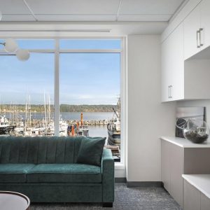 A modern living room with a teal sofa and large window overlooking a marina with boats docked. White cabinets, a wall-mounted TV, and a glass bowl with plants sit on built-in shelves.