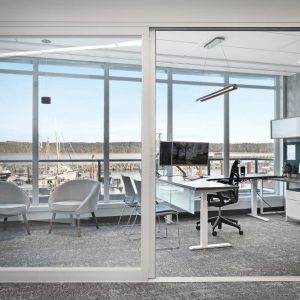 Modern office with glass walls, featuring a desk, office chair, large monitor, cabinets, and seating area with three chairs. Large windows reveal a marina with boats and water in the background.