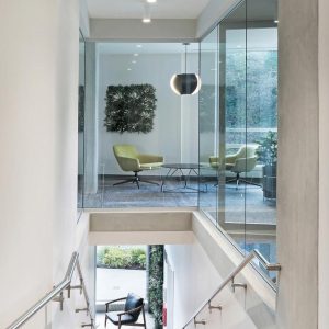 A modern indoor staircase with glass walls leads to a bright lounge area with large windows, light green chairs, a round table, and wall art. An armchair sits at the entrance below, next to greenery outside.