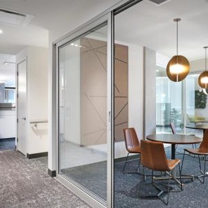 Modern office space with glass-walled meeting room containing a round table and four brown leather chairs, pendant lights overhead, and a blurred figure walking in the hallway outside.