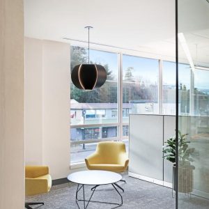 A modern office lounge with two yellow armchairs, a round white coffee table, a black pendant light, a glass wall, and large windows overlooking buildings and trees outside.