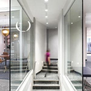 A modern interior hallway with glass walls and a staircase leading downward. A blurred person walks down the steps, and contemporary lighting fixtures are visible in adjacent rooms.
