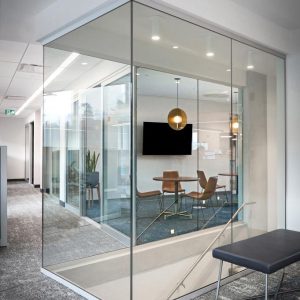 A modern office space with glass walls enclosing a conference room containing a table, chairs, a TV screen, pendant light, and a potted plant. Outside the room, there is a gray bench and carpeted flooring.