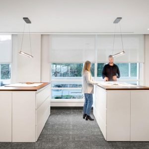 Two people are standing and talking at separate white office workstations in a modern, bright office with large windows and minimalist decor.