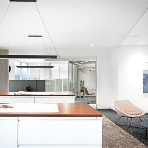 Modern office space with a large wooden counter, two beige chairs around a small black table, abstract wall art, and glass walls revealing another meeting room in the background. Bright and minimal decor.