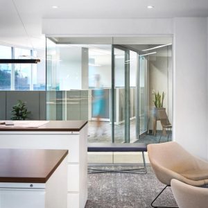 Modern office with white walls, glass meeting room, beige chairs, and a plant. Blurred figure walks behind glass. Natural light enters through windows, creating a bright and clean workspace.