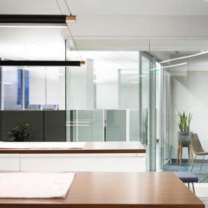 Modern office space with clean lines, glass walls, a long wooden table with blueprints, minimal furniture, indoor plants, and natural light streaming through large windows.