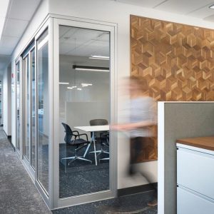 Modern office hallway with a glass-walled meeting room containing three chairs and a table, geometric wood accent wall, and a person walking past cubicles, captured with motion blur.