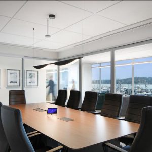A modern conference room with a long wooden table, black chairs, a small tablet on the table, wall art, large windows with a city view, and two blurred figures walking outside the glass walls.