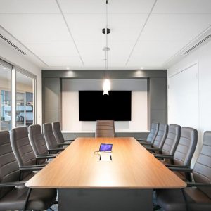 A modern conference room with a long wooden table, ten black leather chairs, a large blank screen on the wall, glass walls on the left, and a tablet placed on the table. The room is well-lit and tidy.