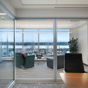 Modern office with glass walls, black chairs, and a wooden table. A person walks past lounge chairs in a seating area. Large windows reveal a marina with boats and water in the background.
