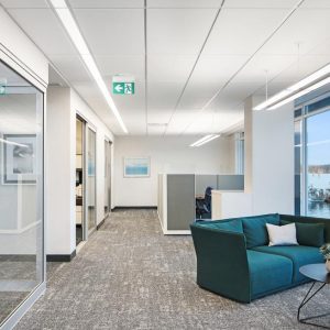 Modern office hallway with glass-walled rooms on the left, a green couch and round coffee table on the right, and large windows overlooking a harbor with boats. The space is bright with overhead lighting.
