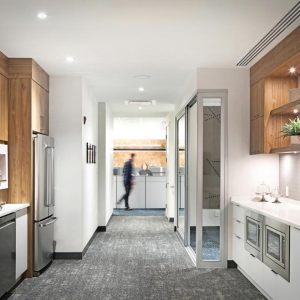 Modern office kitchenette with wood and white cabinetry, built-in appliances, coffee machine, under-cabinet lighting, and plants on shelves. A person is visible walking in the brightly lit background.