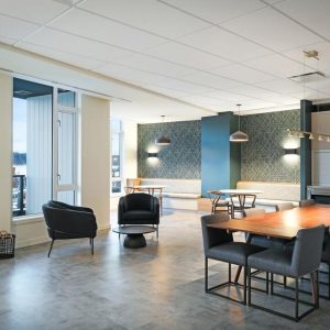 Modern open lounge with large windows, a wooden dining table surrounded by dark chairs, black armchairs, pendant lights, a bench along the wall, and a kitchenette area with shelves and a sink.