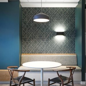 A modern dining nook with a round white table, two wooden chairs with dark seats, a gray cushioned bench, geometric patterned accent wall, and pendant and wall lighting.