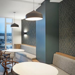Modern café interior with round white tables, wooden chairs, and cushioned bench seating along a wall with geometric-patterned wallpaper. Large windows let in natural light; pendant lights hang from the ceiling.