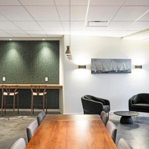 A modern, minimalist lounge with a wooden table, chairs, two armchairs with a small round table, a wall art of sailboats, and a tall counter with two bar stools against a textured green wall.
