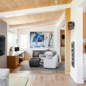Modern living room with light wood floors and ceiling, featuring a cozy seating area with two armchairs, a small desk by the window, abstract wall art, and an open kitchen with wood cabinets.