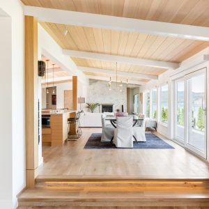 Modern open-concept living and dining area with large windows, wooden floors, light wood ceiling, neutral furnishings, a blue rug, and views of greenery and water outside.