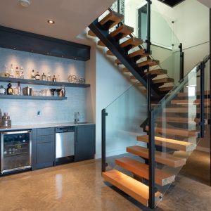 A modern home interior features an open wooden staircase with glass railings. Below the stairs is a sleek bar area with shelves of bottles, a mini fridge, sink, and cabinets on a polished concrete floor.