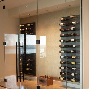 A modern glass-enclosed wine cellar with multiple rows of wine bottles stored horizontally on wall-mounted racks and a wooden crate on the floor, set against a light-colored wall and polished floor.