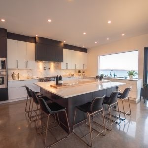 Modern kitchen with a large island, black and white cabinetry, stainless steel appliances, bar stools, marble backsplash, and a large window offering a scenic water view. Sliding doors open to an outdoor patio.