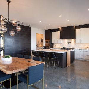 Modern open-concept kitchen and dining area with a wooden dining table, navy chairs, black and white cabinetry, marble backsplash, island with barstools, and contemporary pendant lighting. Plants decorate the bright space.