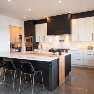 Modern kitchen with white and black cabinets, marble backsplash, a large island with a wooden accent, three black barstools, built-in appliances, potted plants, and recessed lighting.