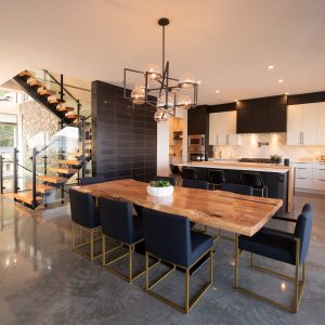 Modern open-concept kitchen and dining area with a large wooden table, eight dark chairs with gold accents, sleek cabinetry, marble countertops, and a glass staircase leading upstairs. Ample natural light fills the space.