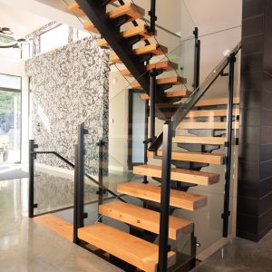 Modern staircase with wooden steps, black metal supports, and glass railings in a bright home with polished concrete floors and patterned wallpaper on one wall.