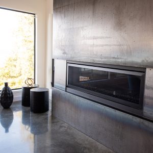Modern living room with a large metal-faced fireplace, polished concrete floor, a window with outdoor view, and two black vases and a small black round table near the corner.