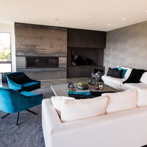 Modern living room with a white sectional sofa, teal accent chairs, a minimalist black fireplace, and textured gray walls. Large windows let in natural light; decor includes throw pillows, a rug, and decorative items on a low table.
