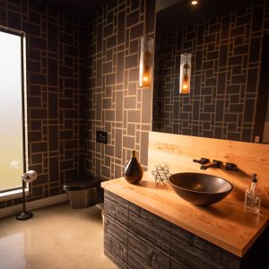A modern bathroom with geometric wallpaper, a black vessel sink on a wooden countertop, pendant lights, and a frosted window. The decor includes a vase, a geometric sculpture, and a soap dispenser.