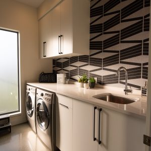 Modern laundry room with white cabinets, a washer and dryer side by side, a sink, potted plants, and bold geometric wallpaper, lit by recessed ceiling lights and natural light from a frosted window.