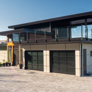 Modern two-story house with large windows, glass balcony railing, flat roof, and two black garage doors, situated on a paved driveway beside a lake on a sunny day.