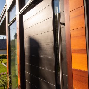 Modern wooden front door with a tall, narrow window and a long vertical handle, set in a house exterior with warm wood paneling and large windows. Sunlight casts shadows across the entryway.