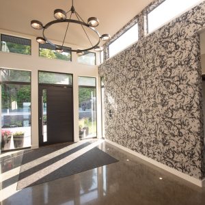 A modern, sunlit entryway with large windows, a dark wooden front door, patterned wallpaper on one wall, polished concrete floors, and a black circular chandelier hanging from the ceiling.