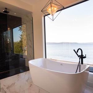 Modern bathroom with a white freestanding bathtub, black faucet, large window overlooking a lake, glass shower, marble floor, and a geometric pendant light. A white robe hangs on the wall.