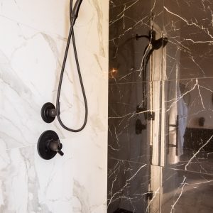 A modern shower features a handheld black showerhead and controls mounted on a wall with white marble tiles, adjacent to a dark, glossy marble wall with a built-in bench.