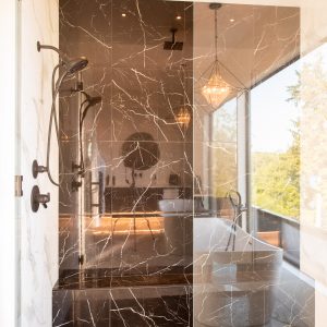 Modern shower with black marble walls and floor, glass door, black fixtures, and a rainfall showerhead. Reflections show a bathtub, large window, and hanging pendant lights outside the shower.