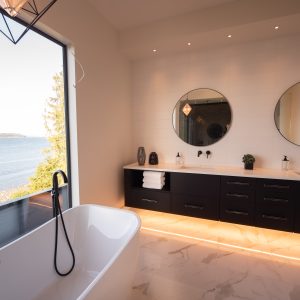 A modern bathroom with a freestanding tub by a large window overlooking a body of water. The room features black cabinets, two round mirrors, and under-cabinet lighting on a marble floor.