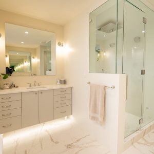 Modern bathroom with a large mirror, beige cabinets, a sink, glass-enclosed shower, towel hanging on a rack, and marble-patterned floor. The room is well-lit and has a clean, minimalist design.