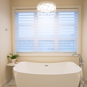 Modern bathroom with a white freestanding bathtub beneath a large window with blinds, a contemporary pendant light above, and a small side table with a plant and candles on marble flooring.