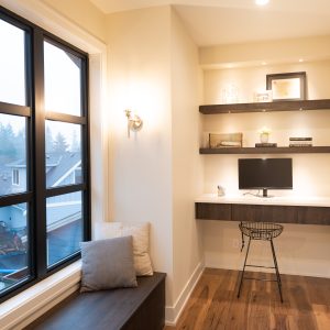 A small, modern home office with a built-in desk, computer monitor, shelves with books and decor, a black wire chair, and a large window overlooking houses and trees. Warm lighting brightens the space.