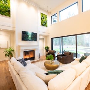 A bright, modern living room with large windows, a white sectional sofa, a fireplace with a TV above it, indoor plants, wooden floors, and greenery art pieces on the walls.
